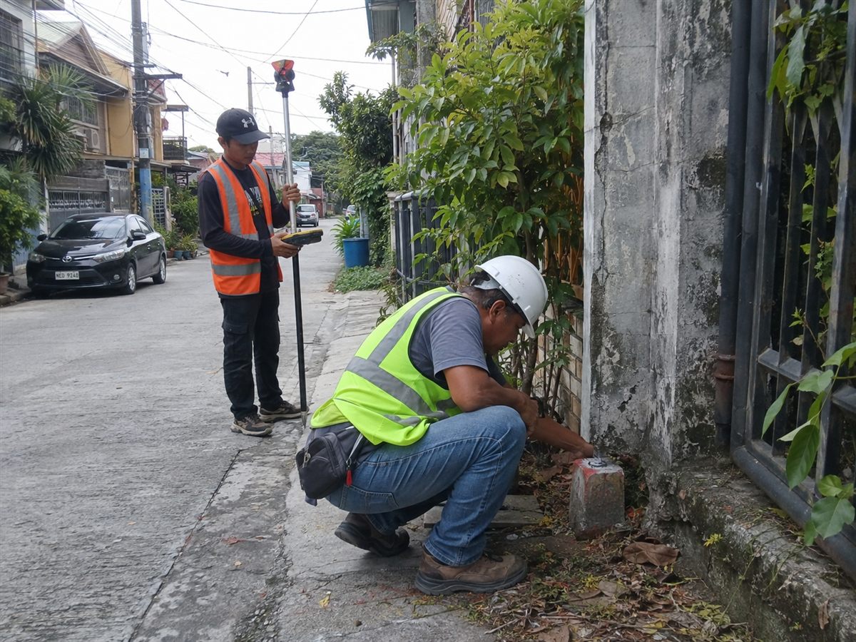 Surveyor in a hard hat crouching to check a painted concrete boundary monument beside a wall while a crew member holds a GNSS rover pole on a residential street