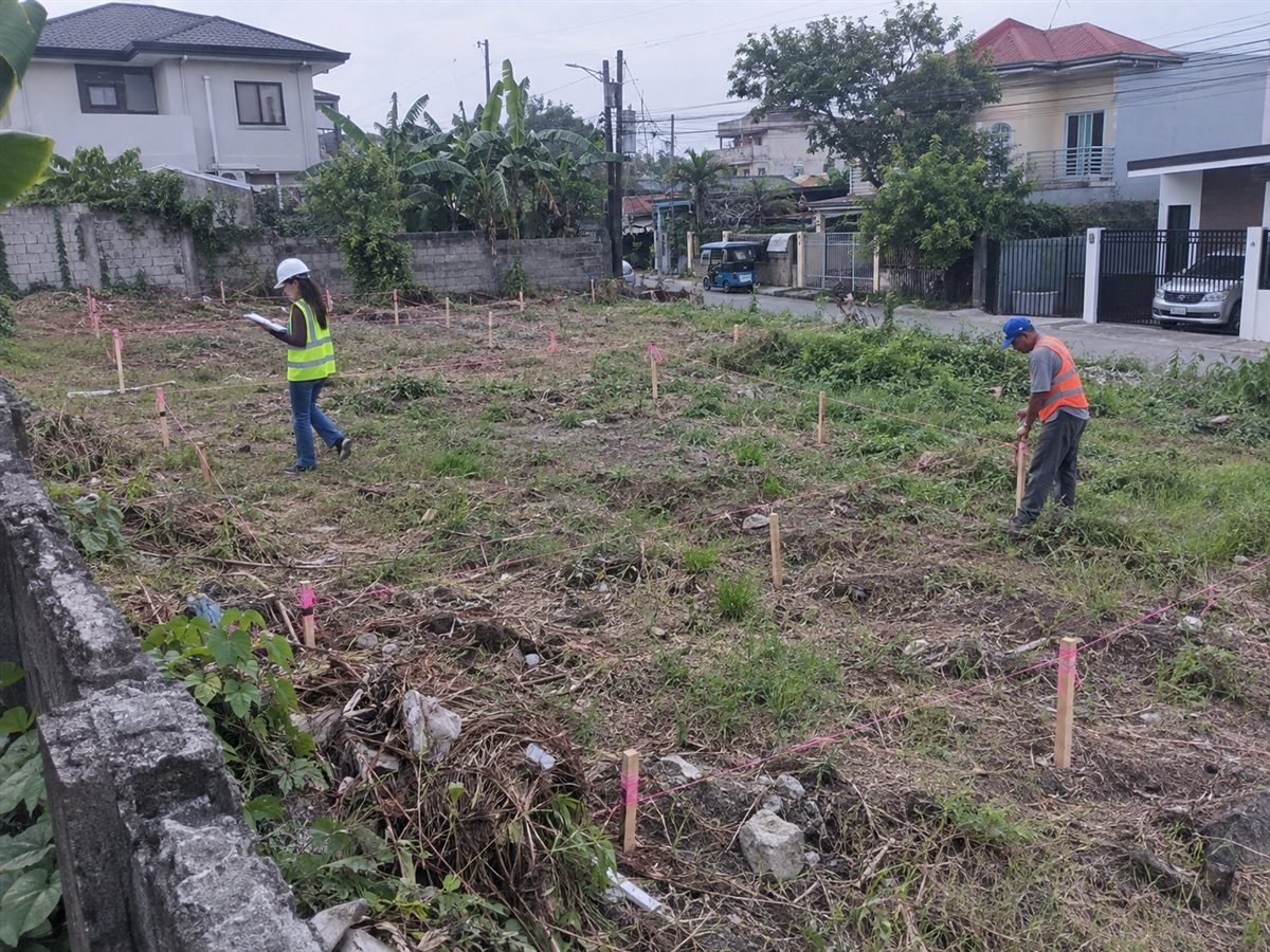 Two surveyors walking a vacant lot marked with wooden stakes and pink string lines to lay out separate parcels, with houses in the background